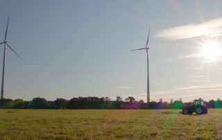 Als Symbolbild für das Projekt Windwende-Videos sieht man ein Foto zweier Windräder in Coesfeld bei Sonnenschein auf einem Feld mit einem Traktor davor.