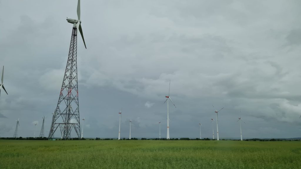 Man sieht auf einer grünen Wiese vor bewölktem, grauen Himmel ein Windrad von Nahem und im Hintergrund 12 weitere Windräder.