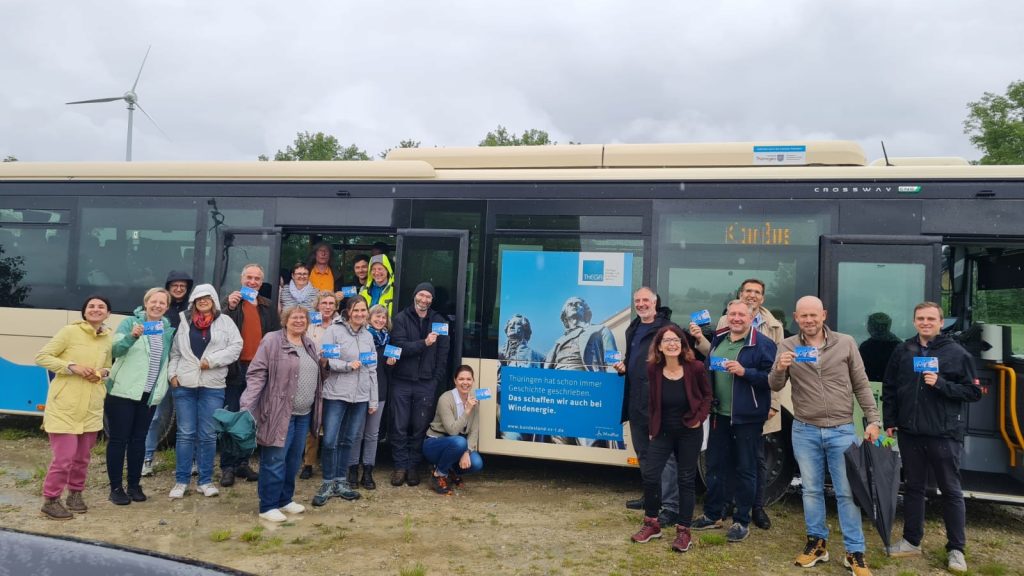 Eine Gruppe von ca. 20 Leuten steht bei trübem Wetter auf einer Wiese vor einem Bus. Im Hintergrund steht ein Windrad. Der Bus trägt das Kampagnen-Plakat mit der Aufschrift "Thüringen hat immer schon Geschichte geschrieben: Das schaffen wir auch bei Windenergie!". Weiterhin ist darauf die Statue von Schiller und Goethe und das Logo der Thüringer Landesenergieagentur und des WindRats zu sehen.