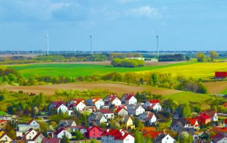 Man sieht eine kleine Stadt im Vordergrund vor weiten Feldern mit blauem Himmel und einigen Windenergieanlagen im Hintergrund.