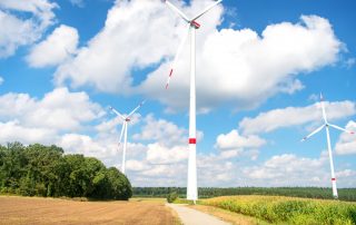 Man sieht zwei Windräder vor blauem Himmel auf einem Feld als Symbolbild für die Beteiligung von Anwohner*innen an Windenergie.