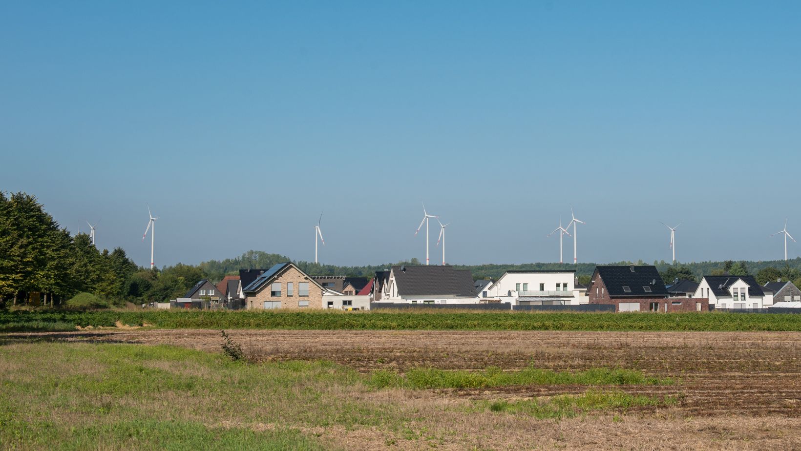 Acker mit kleiner Siedlung vor blauem Himmel mit einigen Windrädern im Hintergrund als Symbol für Umwelt- und Anwohnerschutz bei Windparkplanung.