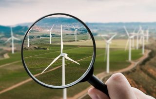 Man sieht Windräder auf einer hügeligen Wiese. Eine Hand hält eine Lupe davor. Symbolbild für "Umweltbundesamt Energiewende"