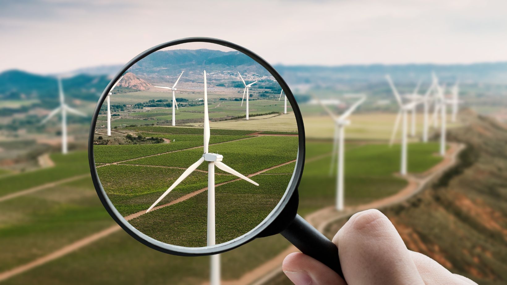 Man sieht Windräder auf einer hügeligen Wiese. Eine Hand hält eine Lupe davor. Symbolbild für "Umweltbundesamt Energiewende"