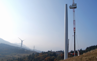 Eine Windenergieanlage im Bau mit Kran in der Natur, vor Bergen mit herbstlicher Bewaldung.
