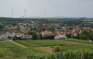 Zu sehen ist ein Dorf. Im VOrdergrund ist ein Weingut zu sehen. Dahinter das Dorf, Wald und Berge. Im Hintergrund einige Windräder.
