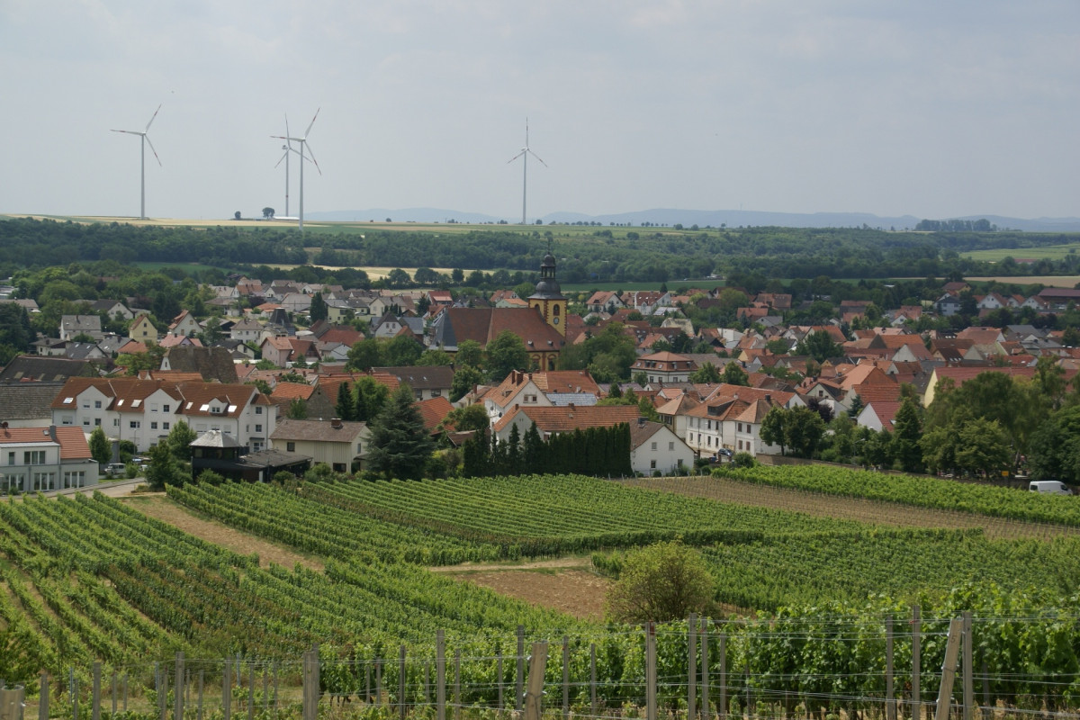 Zu sehen ist ein Dorf. Im VOrdergrund ist ein Weingut zu sehen. Dahinter das Dorf, Wald und Berge. Im Hintergrund einige Windräder.